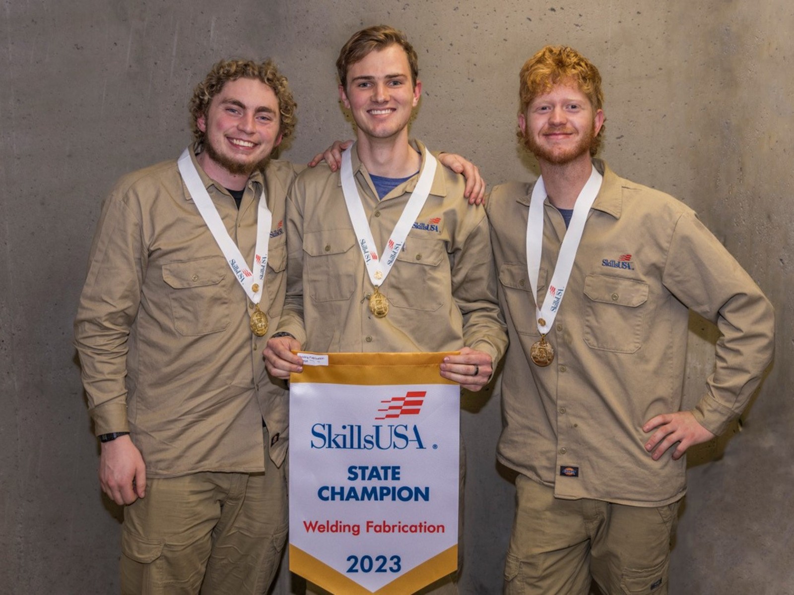 Three men wear gold medals and hold a championship banner.